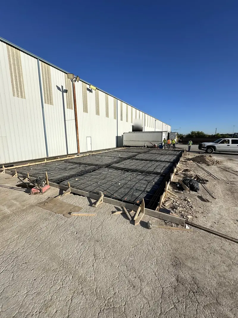 Slab forms and rebar over black plastic beside a long white metal warehouse, crew in safety vests at the far end.