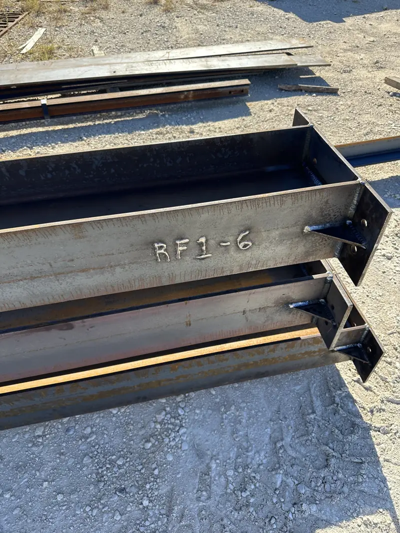 Steel rafter beams with welded end plates, marked RF1-6, laid out on gravel.