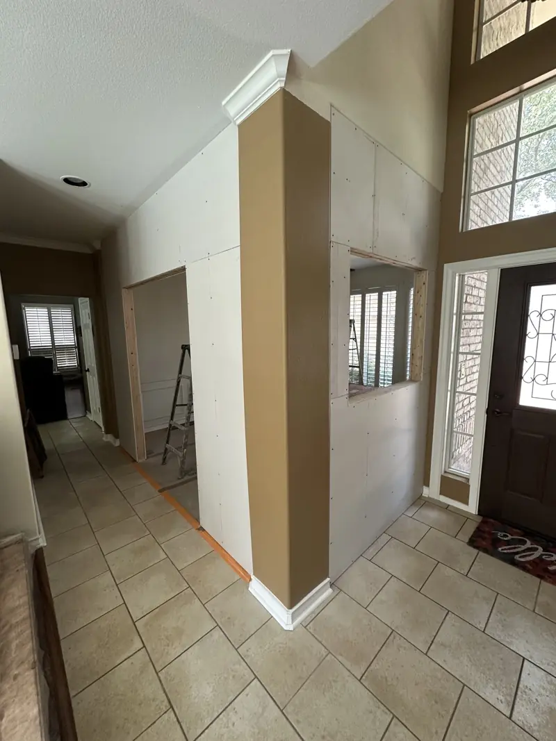 Drywall hung on the new walls seen from the foyer, with a door opening, a window cutout and a painted corner column.
