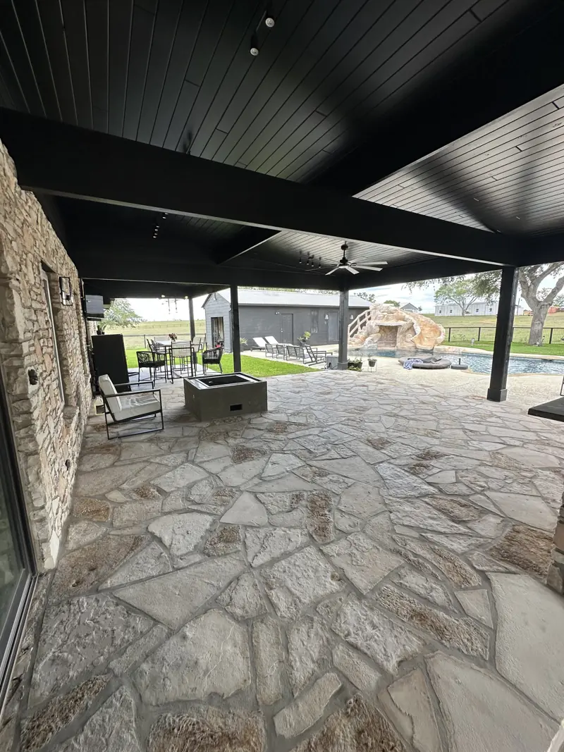 Covered patio with black plank ceiling, ceiling fan, flagstone floor and concrete fire pit, pool and grotto beyond.